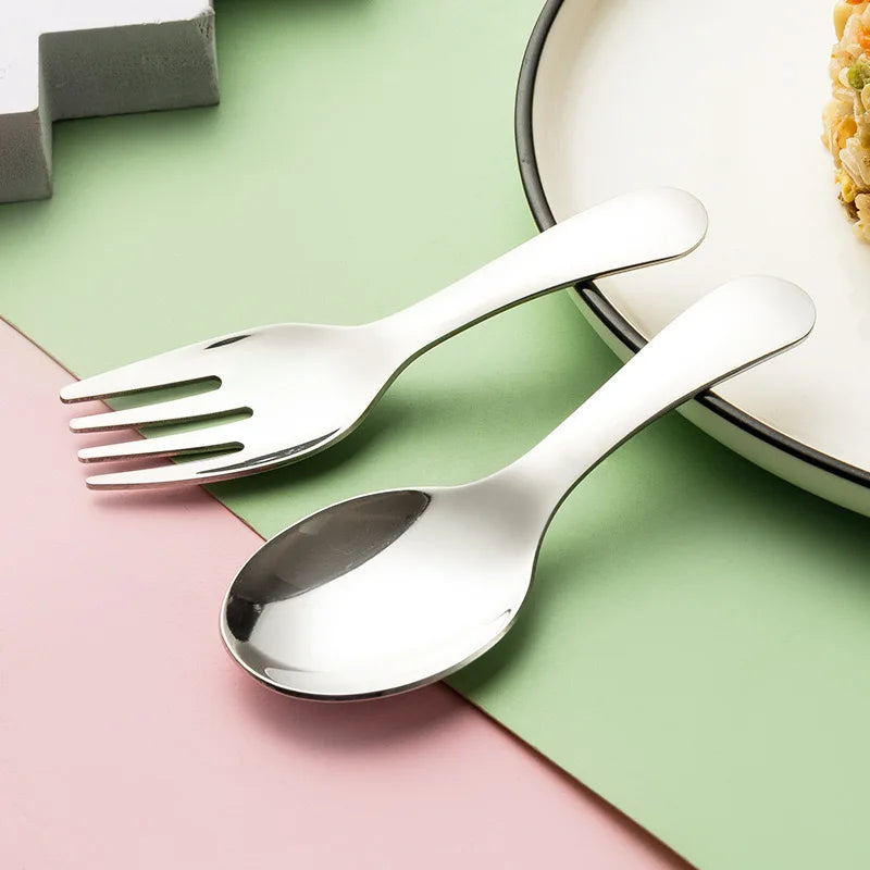 A stainless steel baby spoon and fork set arranged diagonally on a split pastel pink and green background, with a white plate partially visible in the upper right.