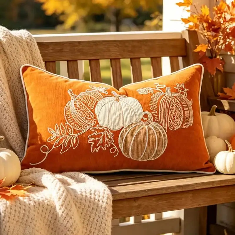 An orange velvet throw pillow with white embroidered pumpkins and leaves, resting on a wooden bench in an autumn outdoor setting with a cream knit blanket and fall decorations.