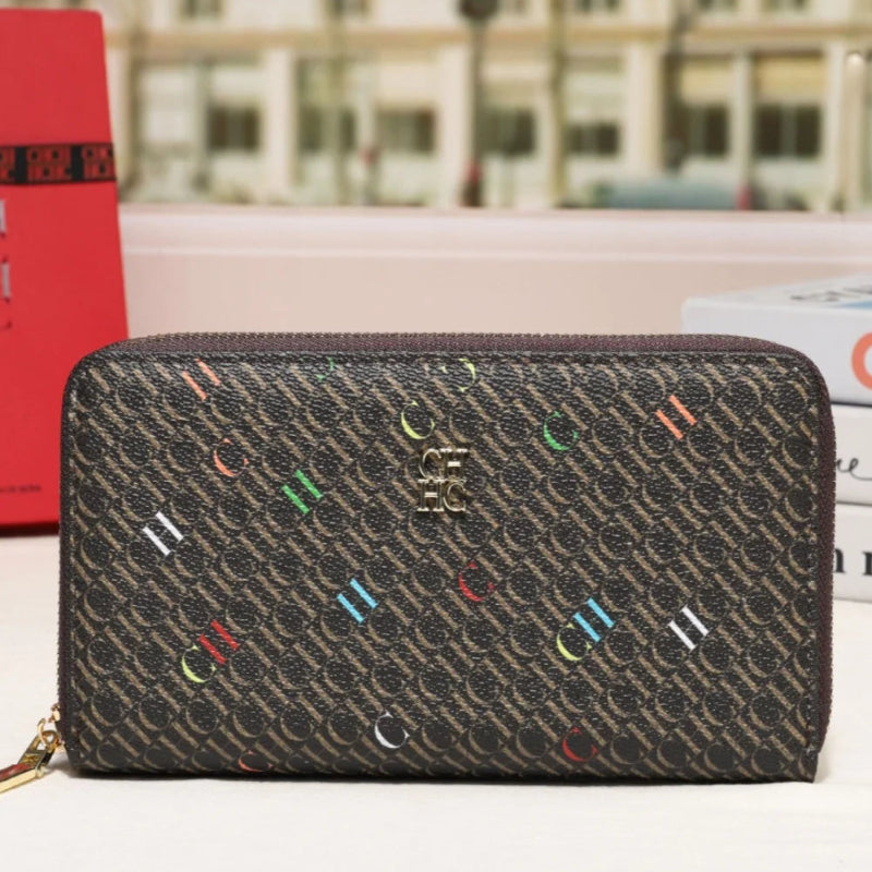 A dark brown monogram zip-around wallet with colorful embroidered letter accents and a gold-tone logo, displayed on a tabletop with a blurred city street background.