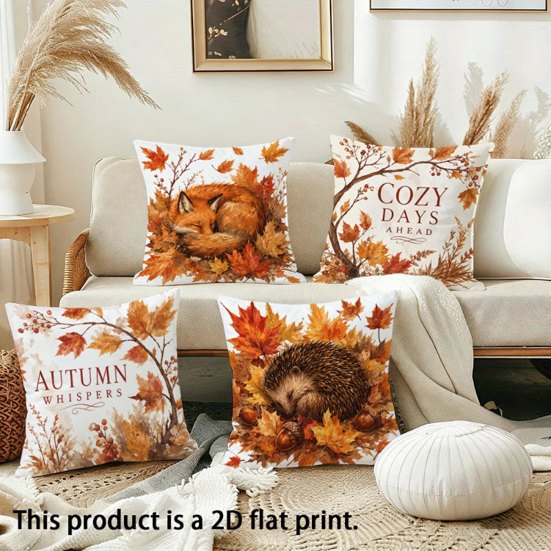 Four decorative throw pillows with autumn-themed 2D flat prints are arranged on a beige sofa in a styled living room. The prints feature a fox, a hedgehog, and text reading 'Autumn Whispers' and 'Cozy Days Ahead' surrounded by orange and yellow leaves.