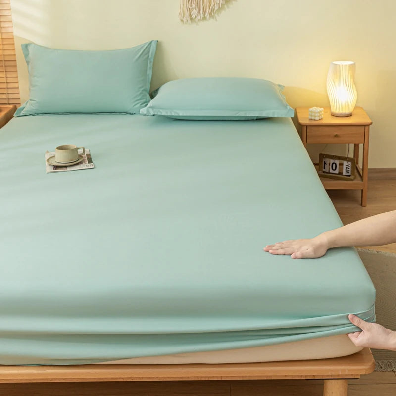 A light mint green bed sheet set with two matching pillowcases on a wooden bed frame, shown in a cozy bedroom setting with a hand stretching the sheet to demonstrate its elasticity.