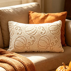 A cream-colored textured lumbar pillow with a raised pumpkin pattern, styled on a beige sofa next to an orange throw pillow and a chunky knit blanket.