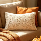 A cream-colored textured lumbar pillow with a raised pumpkin pattern, styled on a beige sofa next to an orange throw pillow and a chunky knit blanket.