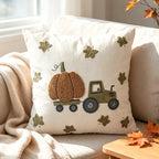 A square cream throw pillow with an autumn-themed embroidered design of a pumpkin on a tractor and scattered maple leaves, styled on a beige sofa with a blanket and fall leaves.