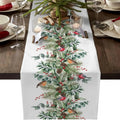 A white table runner printed with a festive Christmas design featuring green pine branches, red berries, pinecones, and small birds, displayed on a dark wood table with place settings and candles.