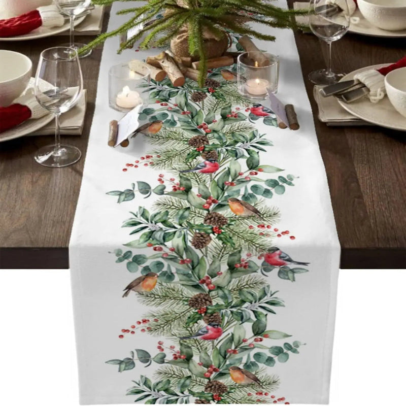 A white table runner printed with a festive Christmas design featuring green pine branches, red berries, pinecones, and small birds, displayed on a dark wood table with place settings and candles.