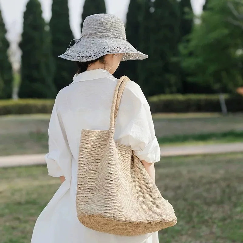 A woman viewed from behind wearing a white shirt and a wide-brim straw hat, carrying a large woven straw tote bag over her shoulder in an outdoor park setting.