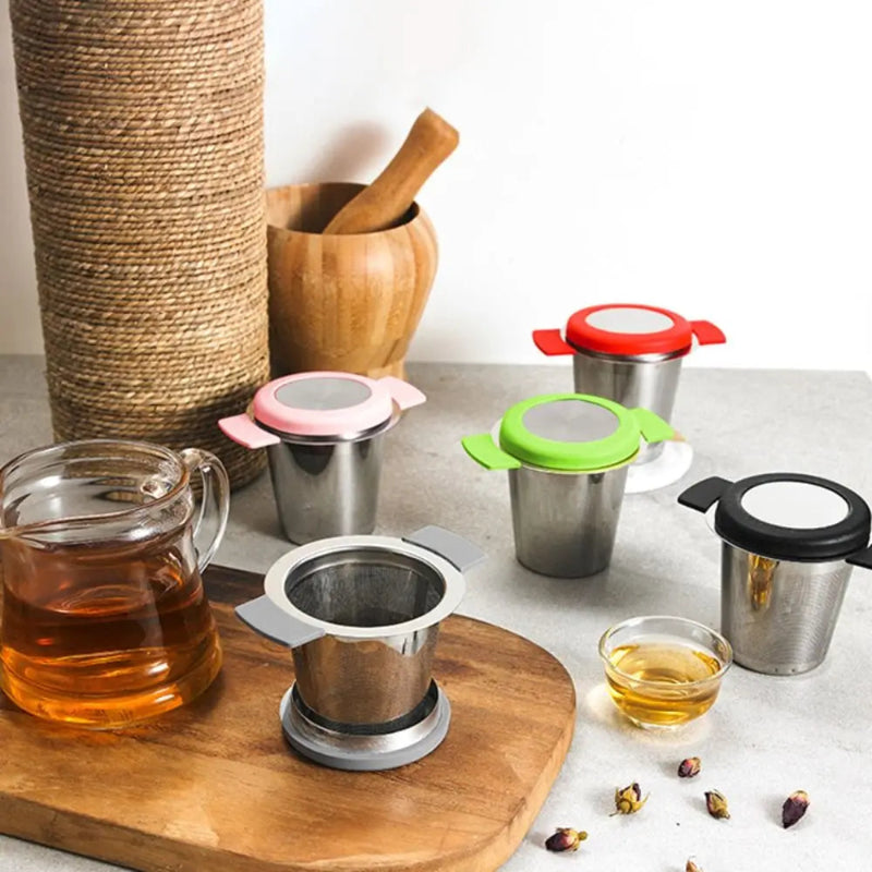 A set of stainless steel tea infusers with colorful lids and handles in pink, green, red, and black, displayed on a wooden cutting board with a glass teapot of tea and dried rose petals.