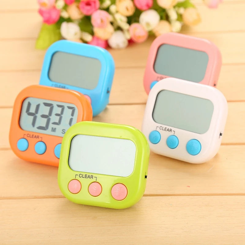 Four small digital countdown timers in orange, blue, pink, and green, arranged on a light wood surface with blurred flowers in the background.