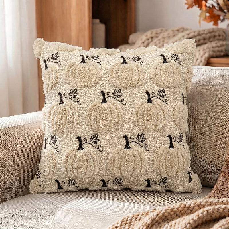 A square decorative throw pillow with a textured cream background and black embroidered pumpkins, styled on a beige sofa with a cozy knit blanket.