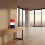 A small rectangular wristlet or clutch with a red, white, and navy color-blocked design and a red wrist strap, placed on a light wood console in a modern high-rise room with large windows overlooking a city skyline.