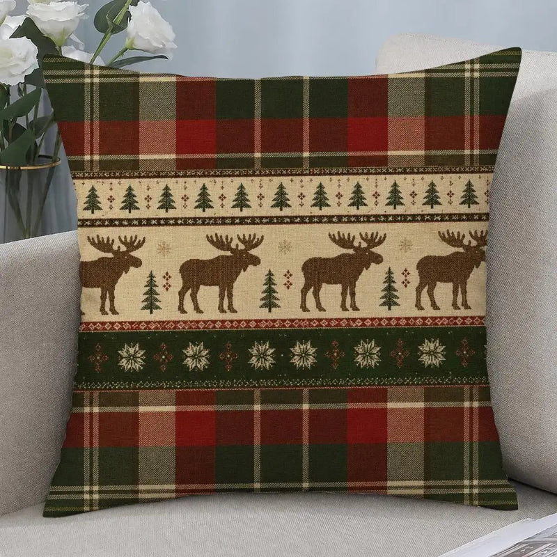 A square throw pillow with a festive Christmas pattern featuring moose silhouettes, pine trees, snowflakes, and plaid borders in red, green, beige, and brown, resting on a light gray sofa.
