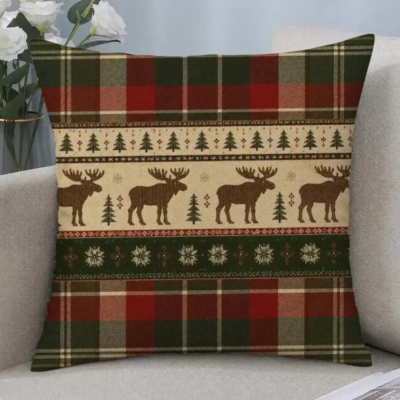 A square throw pillow with a festive Christmas pattern featuring moose silhouettes, pine trees, snowflakes, and plaid borders in red, green, beige, and brown, resting on a light gray sofa.