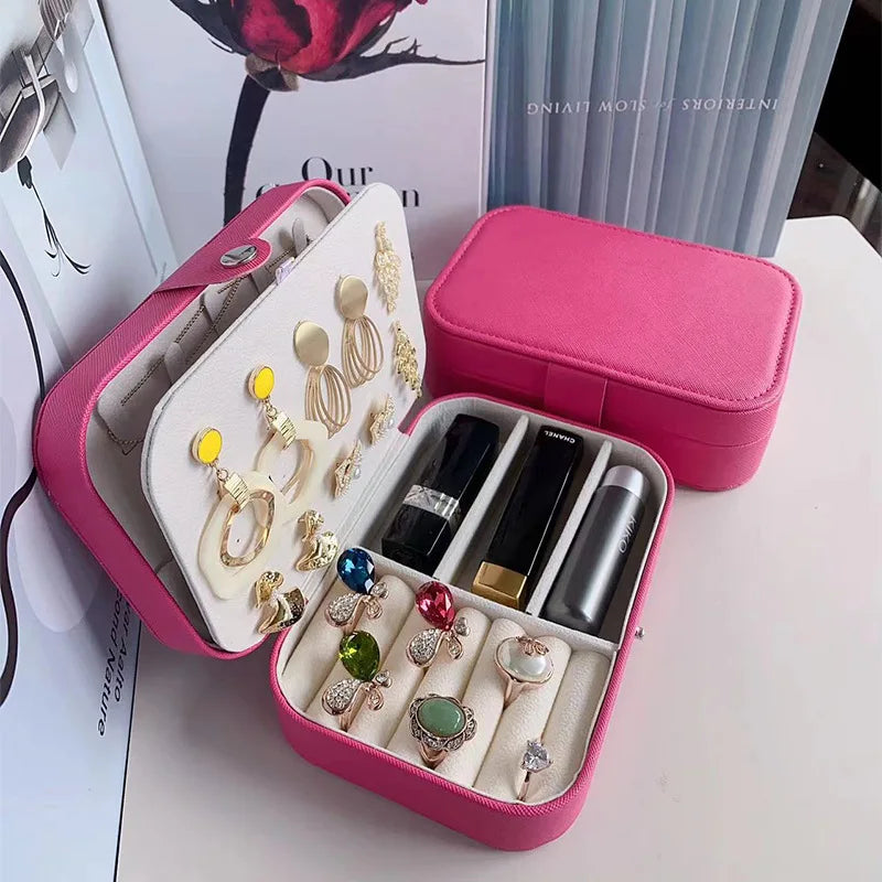 A pink hard-shell ring and jewelry organizer shown open and closed on a tabletop. The open case contains multiple compartments holding rings, earrings, and small accessories, with a cream insert lining.