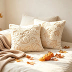 Two cream throw pillows with autumn-themed embroidery, one featuring pumpkins and the other maple leaves, styled on a bed with a knit blanket, small pumpkins, and scattered fall leaves.