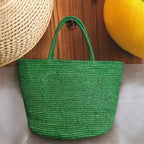 A green woven straw tote bag with two top handles, hanging against a wooden background with a straw hat and a yellow citrus fruit partially visible in the frame.