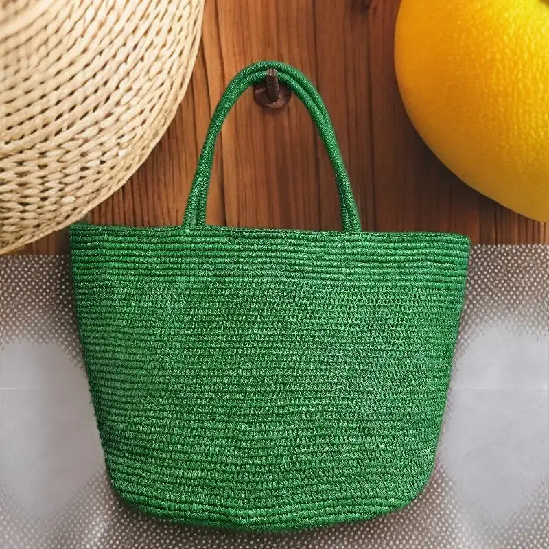 A green woven straw tote bag with two top handles, hanging against a wooden background with a straw hat and a yellow citrus fruit partially visible in the frame.