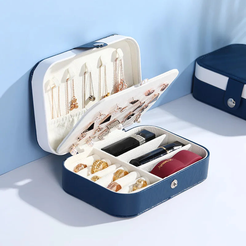 A navy blue and white jewelry organizer case shown open, containing rings, necklaces, and other jewelry, with a matching closed case partially visible in the background against a light blue wall.