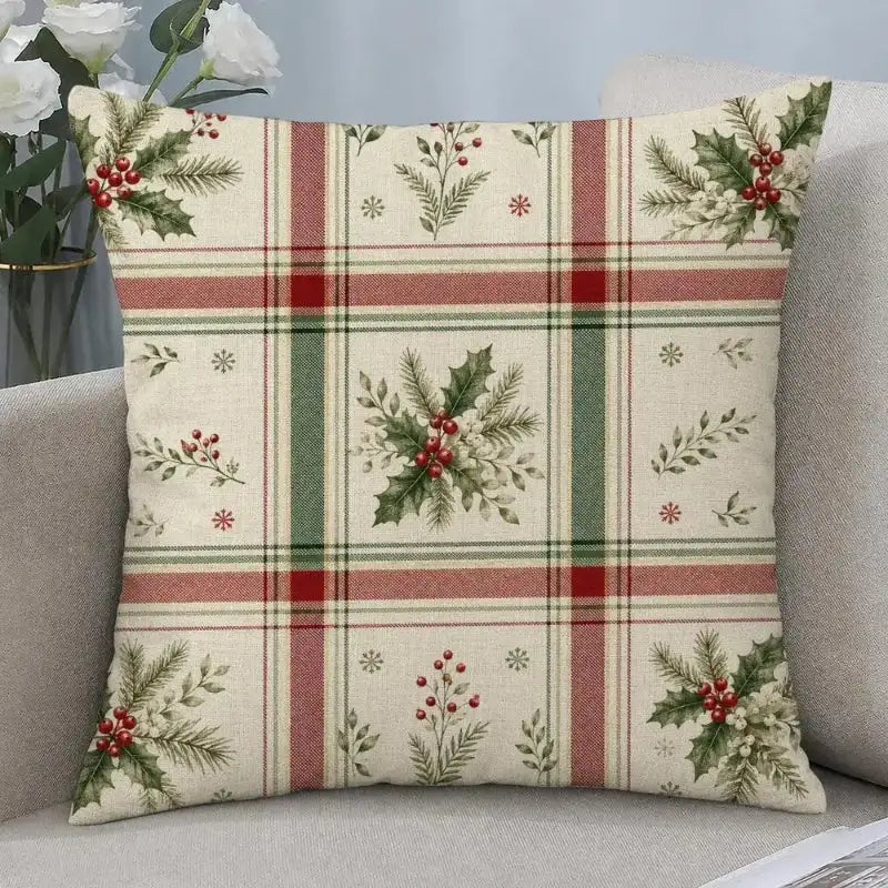 A square throw pillow with a Christmas-themed plaid pattern featuring holly leaves, pine branches, and red berries, resting on a light gray sofa.