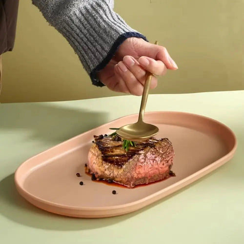 A close-up of a hand holding a gold spoon over a grilled steak on a peach-colored oval plate, set against a soft beige background.