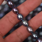 A close-up of a hand holding a strand of dark, iridescent baroque pearls. The pearls have a deep purple and blue-gray color with a glossy, metallic sheen.