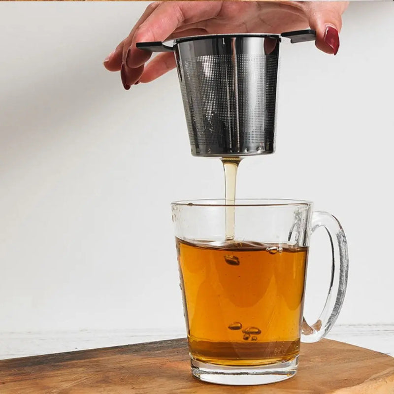 A hand with dark red painted nails holds a small stainless steel mesh tea infuser above a clear glass mug filled with amber tea, with tea dripping into the cup. The mug sits on a wooden surface against a plain light background.