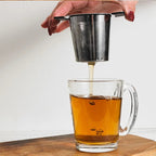 A hand with dark red painted nails holds a small stainless steel mesh tea infuser above a clear glass mug filled with amber tea, with tea dripping into the cup. The mug sits on a wooden surface against a plain light background.