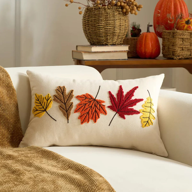 Decorative pillow with embroidered autumn leaves on a couch, with pumpkins and books in the background.