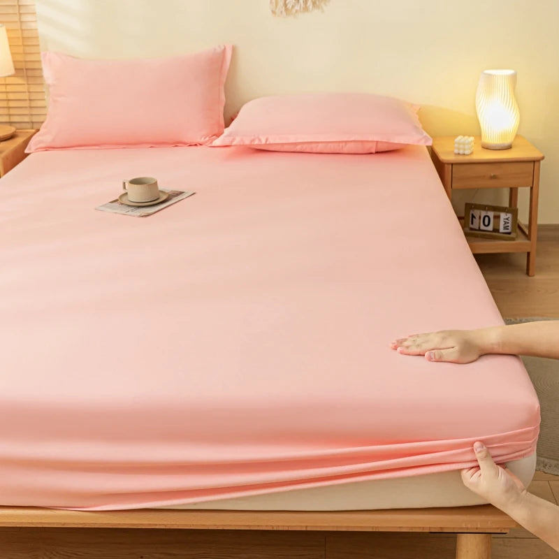 A pink fitted sheet and matching pillowcases on a wooden bed in a bedroom setting, with a person's hands pulling the sheet down over the mattress.