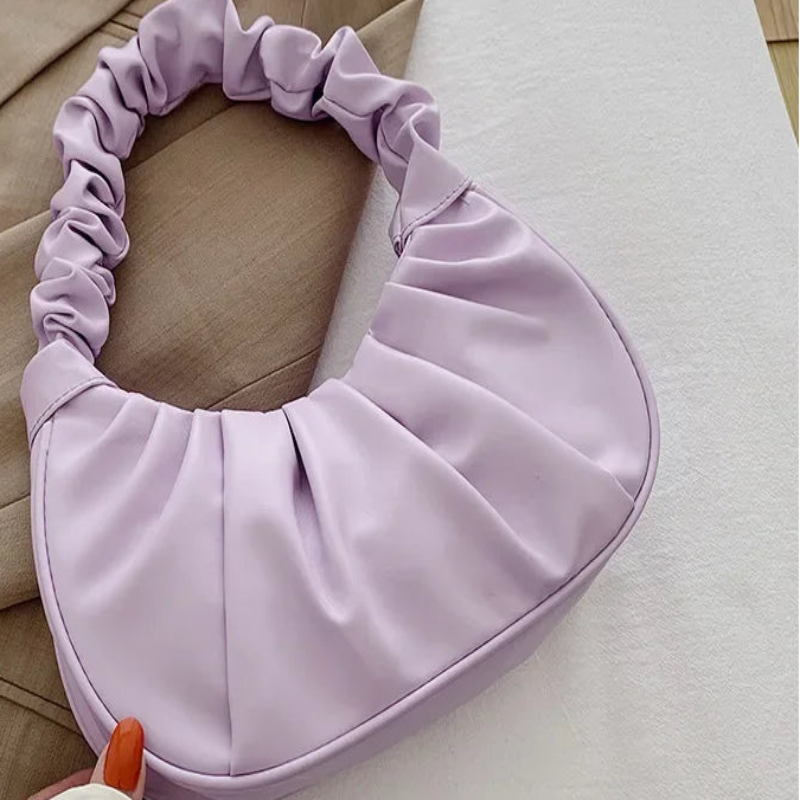 A light purple crescent-shaped shoulder bag with a gathered, ruched handle, styled on a beige blazer over a white fabric background.