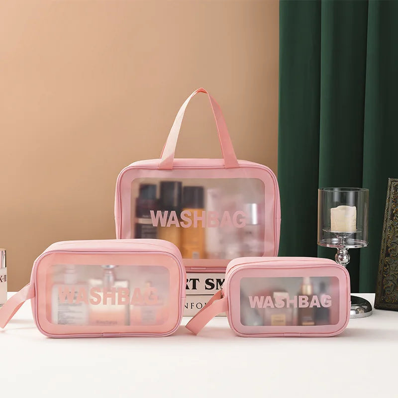 Three pink translucent cosmetic bags of different sizes with top handles, arranged on a tabletop against a beige wall and dark green curtain. The bags have clear front panels with the word 'WASHBAG' printed in pink and contain assorted bottles and jars.