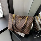 A brown monogram handbag with gold-tone chain straps and a deer charm, displayed on a dark table next to a mirror reflecting its side.