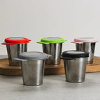Four stainless steel travel mugs with different colored lids (red, green, pink, and gray/black) arranged on a wooden board against a neutral light gray background.