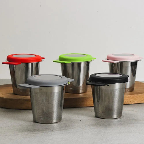 Four stainless steel travel mugs with different colored lids (red, green, pink, and gray/black) arranged on a wooden board against a neutral light gray background.