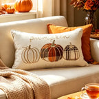 A cream-colored rectangular throw pillow with embroidered autumn pumpkins and the word 'pumpkin' in script, styled on a beige sofa with an orange accent pillow, a knitted blanket, and fall decor in a warm living room setting.