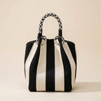 A black and off-white vertically striped bucket bag with braided top handles and silver-tone hardware, photographed against a solid beige background.