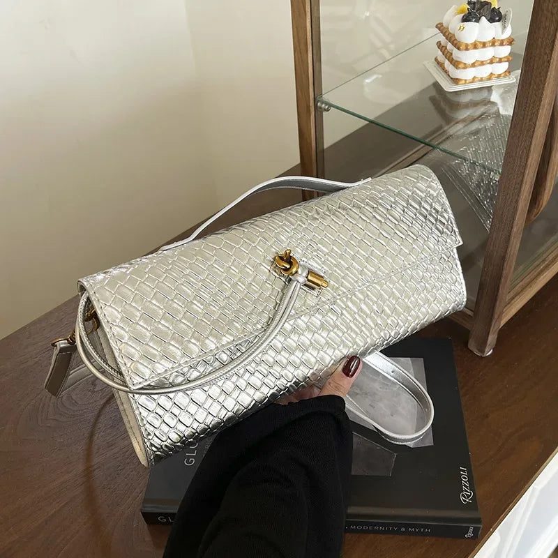 Silver textured handbag held by a person on a wooden surface with a glass cabinet in the background.