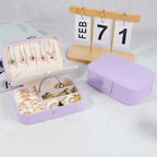 A lavender jewelry organizer box shown open and closed on a white surface, with rings and earrings displayed inside. A wooden calendar stand reading FEB 7 1 and part of a book are visible in the background.