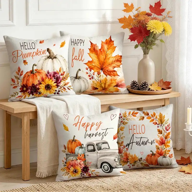 Four decorative throw pillows with autumn-themed prints are arranged on a wooden bench and floor in a styled room. The pillows feature fall leaves, pumpkins, sunflowers, and phrases like 'Hello Pumpkin,' 'Happy fall,' and 'Happy harvest.