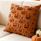 An orange-brown textured throw pillow with raised jack-o'-lantern faces and fringe edges, styled on a cream couch next to small pumpkins.