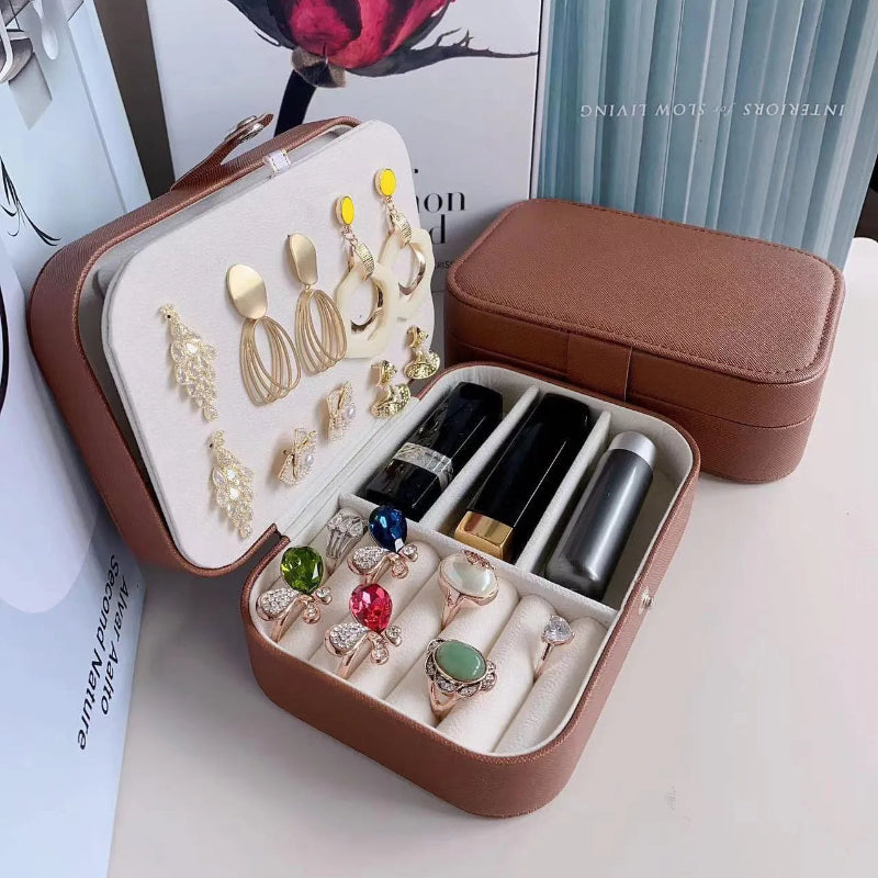 A brown faux-leather jewelry organizer case shown open, containing rings, earrings, and lipsticks, with the closed case placed behind it on a tabletop.