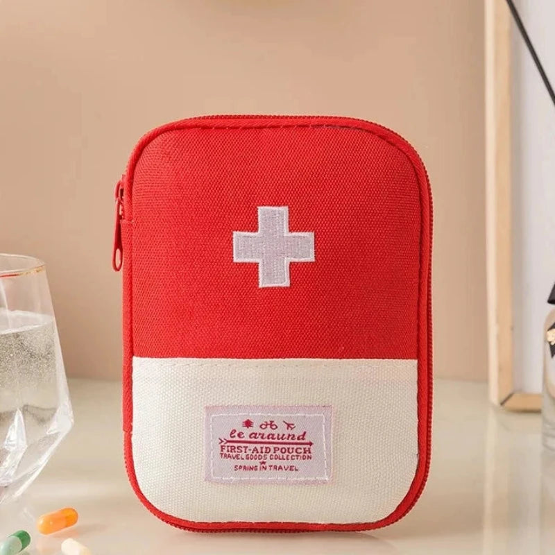 A red and white first aid pouch with a white cross on the front, displayed on a tabletop with a glass of water and small pills nearby. Chinese text at the bottom indicates dimensions: 10.