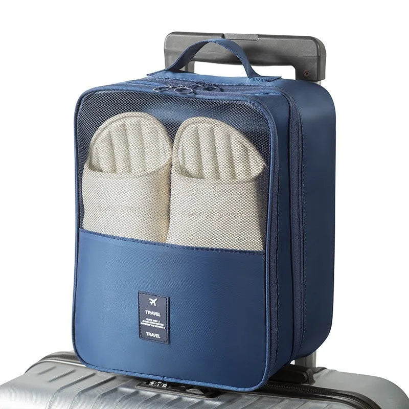 A blue travel shoe bag with a mesh front window showing a pair of off-white slippers inside, placed on top of a silver suitcase against a white background.