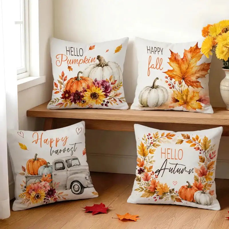 Four decorative throw pillows with autumn-themed prints are arranged on a wooden bench and floor in a bright room. The pillows feature fall illustrations including pumpkins, leaves, flowers, and phrases such as 'Hello Pumpkin,' 'Happy fall,' 'Happy harvest,' and 'Hello Autumn.