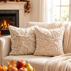 Two cream textured throw pillows with embroidered autumn motifs, styled on a beige sofa with a chunky knit blanket, a fireplace in the background, and fall decor in the foreground.
