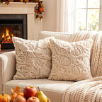 Two cream textured throw pillows with embroidered autumn motifs, styled on a beige sofa with a chunky knit blanket, a fireplace in the background, and fall decor in the foreground.
