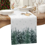 A white table runner with a snowy winter forest design, draped over a wooden dining table set with white plates, silverware, and glassware.