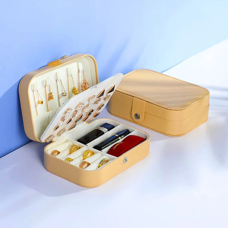 A tan rectangular jewelry organizer box shown open and closed on a white surface against a blue background. The open box displays compartments holding earrings, rings, and a pen, with a red velvet-like insert visible inside.