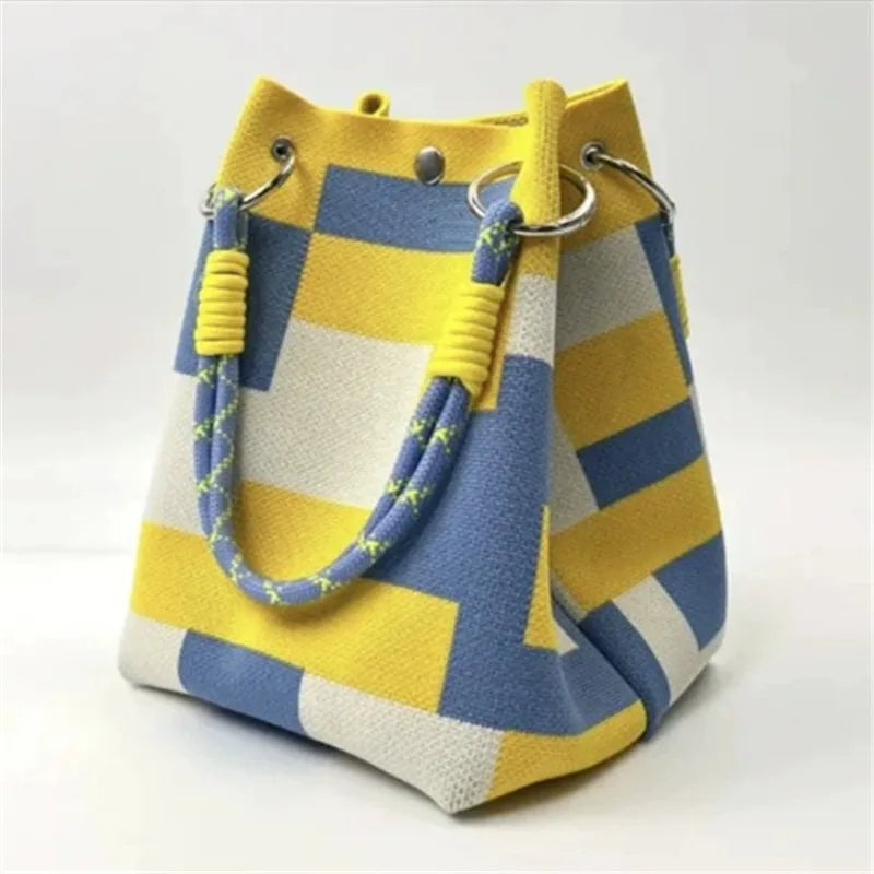 A bucket-style handbag with a bold geometric checkered pattern in yellow, blue, and off-white. It features thick woven rope handles with yellow accents and silver-tone metal hardware.