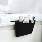Black bed caddy holding a phone, book, and remote control on a white bed.